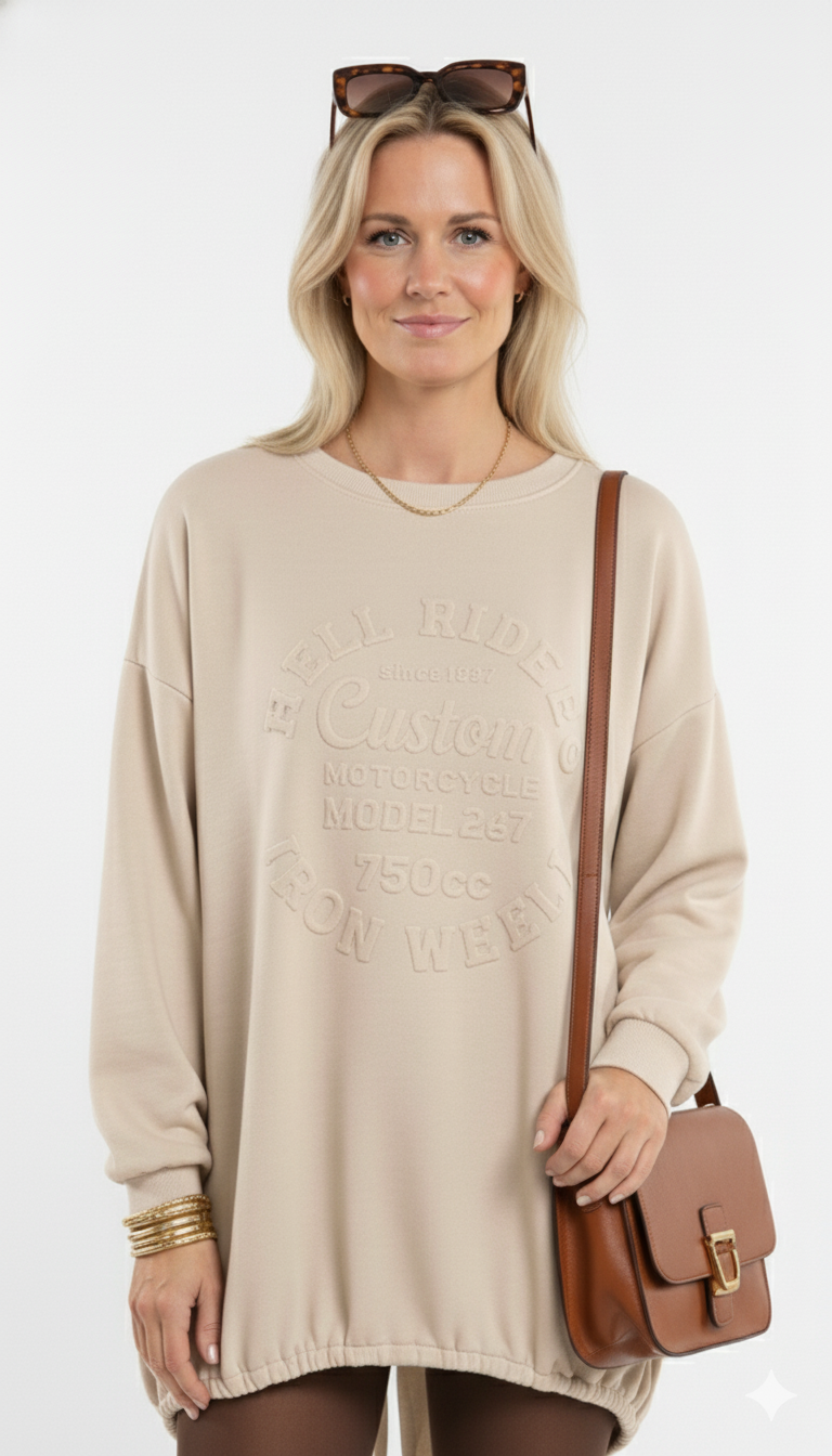 Vrouw draagt beige oversized sweaterdress met reliëfprint ‘Custom Motorcycle’, gecombineerd met bruine tas en gouden accessoires – casual en stijlvolle look.