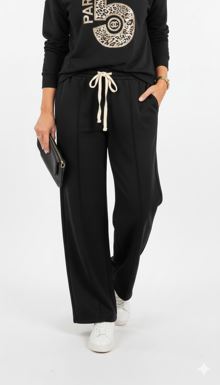 Vrouw draagt zwarte wide leg broek met strikceintuur, gecombineerd met witte sneakers en clutch.