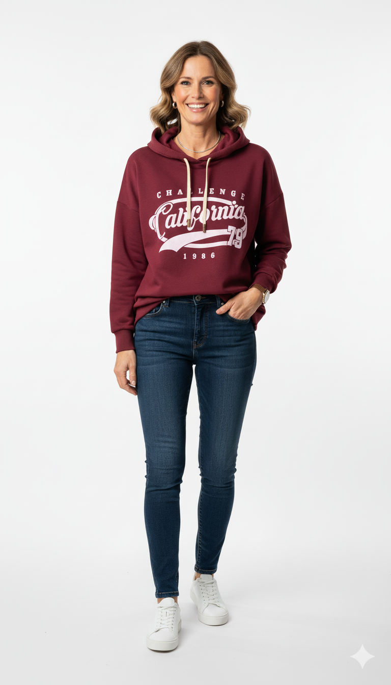 Vrouw met bordeaux hoodie met ‘California Challenge 1986’ print, jeans en witte sneakers – sportieve damestrui met vintage opdruk.