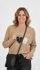 Vrouw draagt camel trui met ‘Steal My Heart’-print en hartjesmotief, gecombineerd met zwarte jeans en schoudertas.