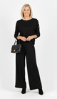 Vrouw draagt zwarte dames set met gouden knopen op de schouders en rechte broek – elegante co-ord outfit met zwarte tas en enkellaarsjes.