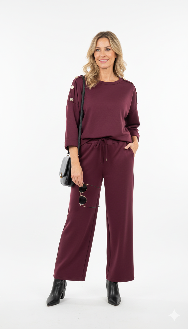 Vrouw draagt bordeaux dames set met gouden knopen op de schouders en rechte broek elegante co-ord outfit met zwarte enkellaarsjes en tas.