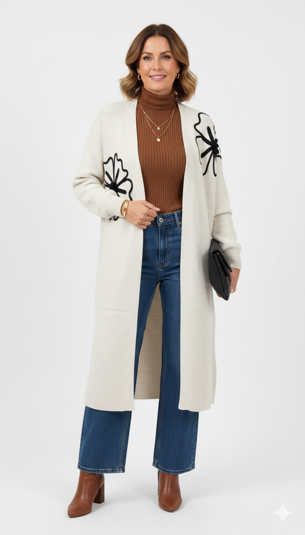 Model draagt een crèmekleurig lang vest met zwarte bloemenapplicatie van Blushfashionstore, gecombineerd met een bruine coltrui, jeans en bruine enkellaarsjes.