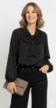 Model draagt een elegante zwarte blouse met strik en gouden stippen van Blushfashionstore, gecombineerd met een zwarte broek.