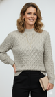 Model draagt een beige kabeltrui met ronde hals van Blushfashionstore, gecombineerd met een zwarte broek en gouden sieraden.